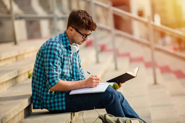 年轻的学生戴着眼镜站在学校或大学的楼梯上 看着他的作业 读一本书 做笔记 — 图库照片