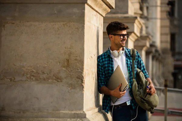 young student with glasses and backpack, leaning with his laptop in hands on colmn of university or school, smiling