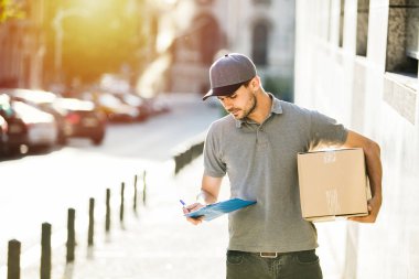 Senin nakliye işte. teslim etmek teslimatçı sokakta için doğru adres arıyor onun karton kutu ile kap ayakta ile gri gömlek