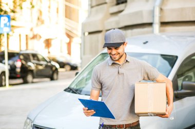 Senin nakliye işte. arabasını, önünde son ayrıntılar için formdaki dışında sokakta arıyorum onun karton kutu ile kap ayakta ile gri tişörtlü teslimat adam