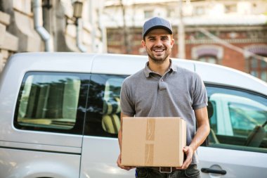 Senin nakliye işte. sokakta teslim arabasının önünde gülümseme ile kameraya bakarak onun karton kutu ile kap ayakta ile gri tişörtlü adam mutlu teslim