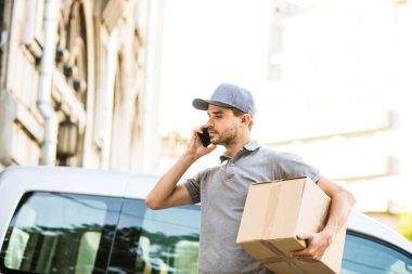 Senin nakliye işte. teslimatçı sokak paketi teslim etmesi için istemci arama ve tarafına bakarak onun karton kutu ile kap ayakta ile gri gömlek