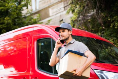 Senin nakliye işte. teslimatçı sokak paketi teslim etmesi için istemci arama ve tarafına bakarak onun karton kutu ile kap ayakta ile gri gömlek