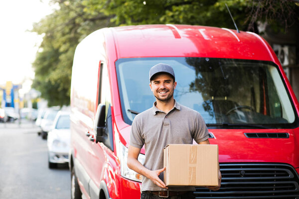 your shipping is here. happy delivery man in grey shirt with cap standing with his cardboard box on the street looking to camera with smile, in front of his delivery car
