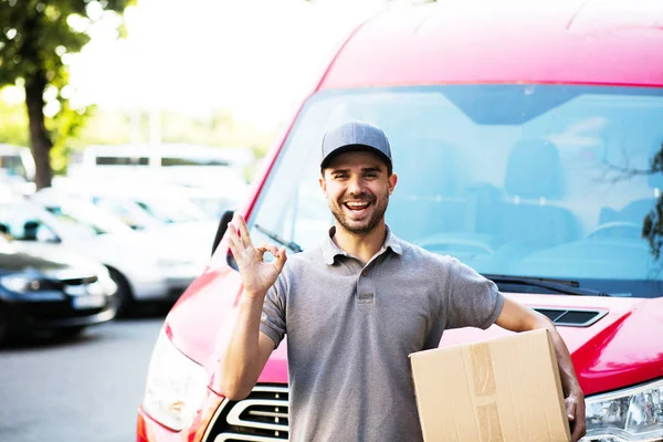 your shipping is here. happy delivery man in grey shirt with cap standing with his cardboard box on the street looking to camera with smile showing confidence, in front of his delivery car