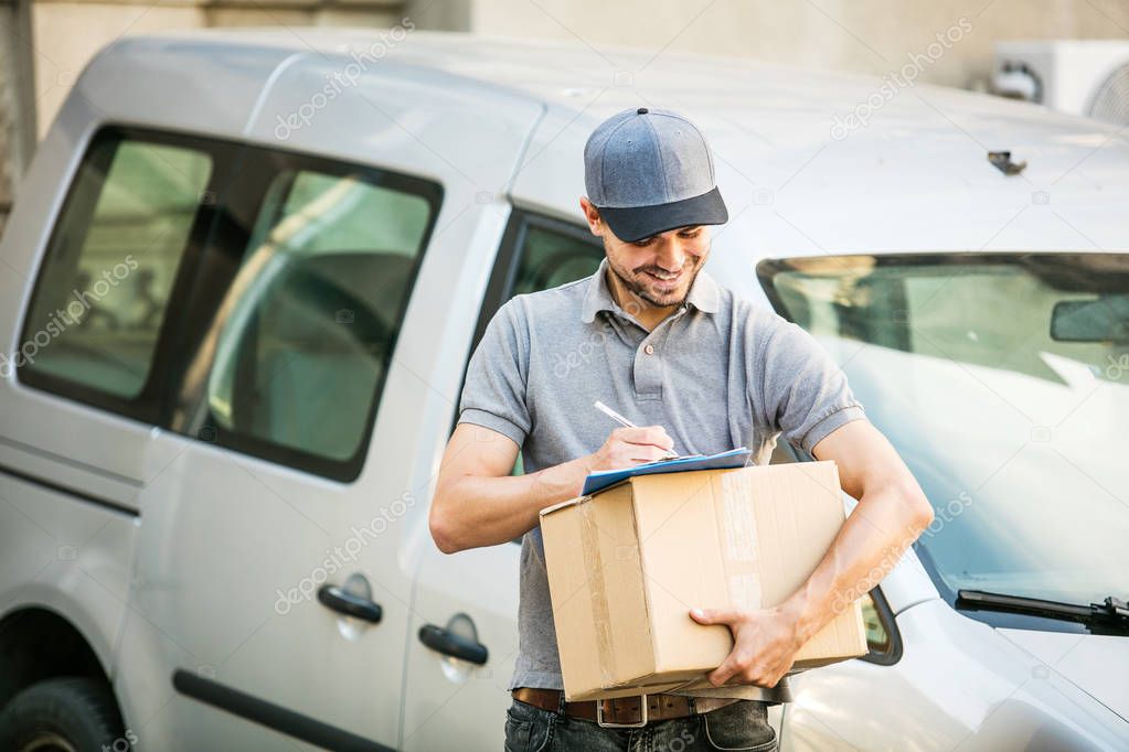 your shipping is here. young delivery man in grey shirt with cap standing with his cardboard box completing the form for deliver, ready to meet the client, outside on the street in front of his car