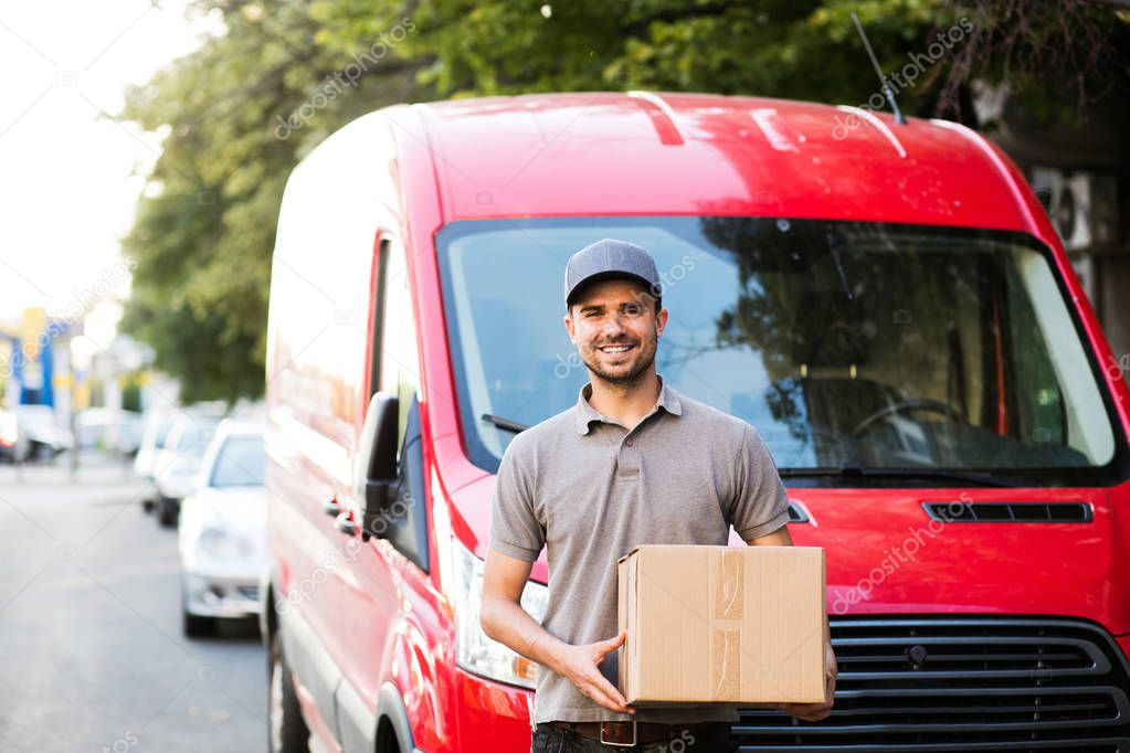 your shipping is here. happy delivery man in grey shirt with cap standing with his cardboard box on the street looking to camera with smile, in front of his delivery car