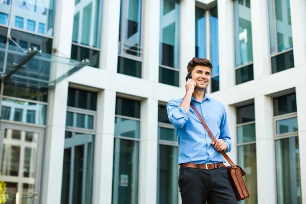 confident and successful antrepreneur, office man or corporate guy, standing with his leather bag and elegant outfit in front of an office building and talking at cellphone