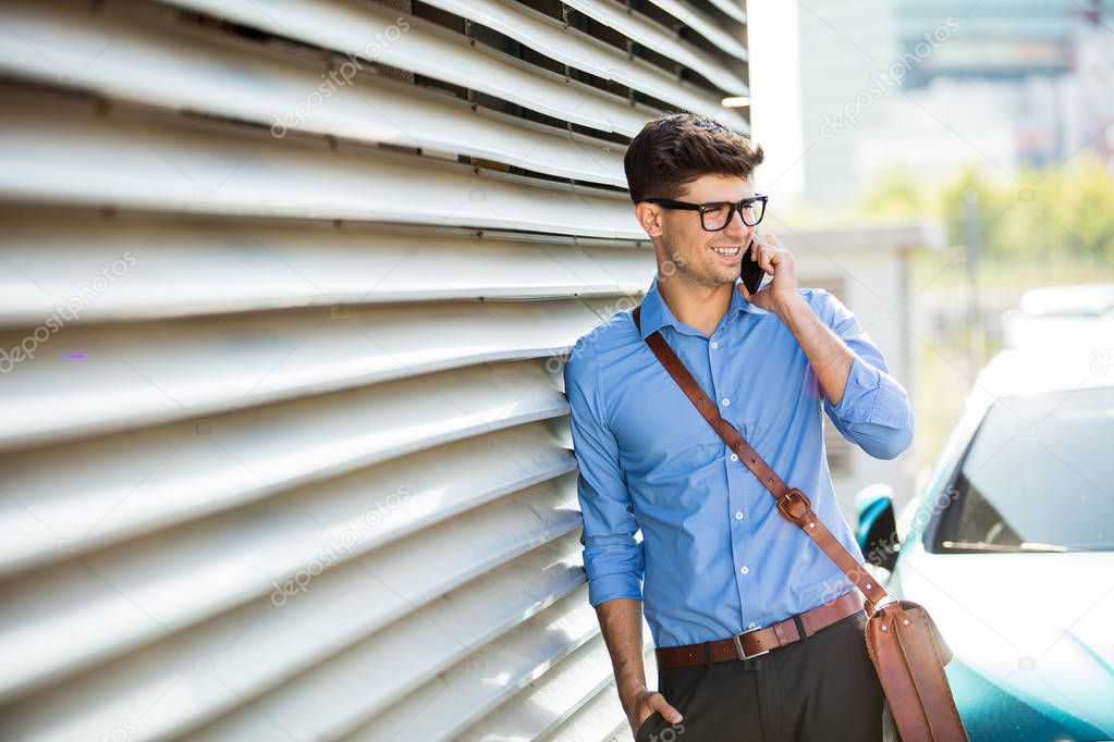 successful antrepreneur, office man, with glasses standing against a wall with sun behind, using his cellphone