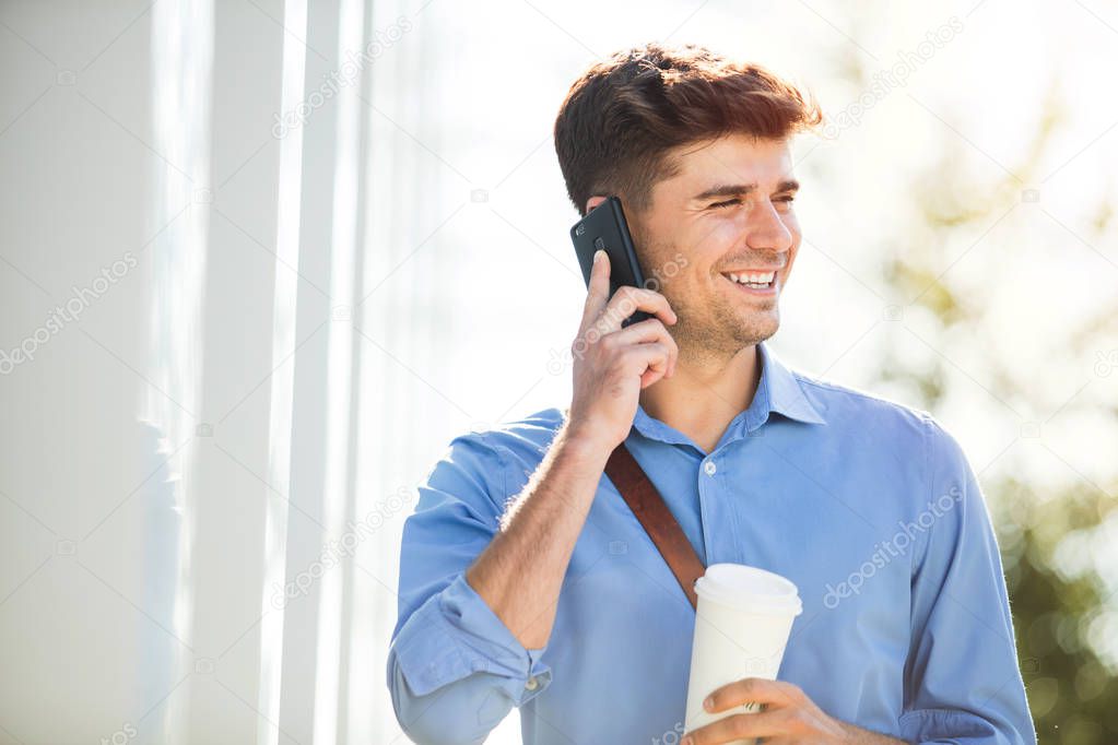 handsome young man going to work, at the office, in blue shirt with coffee in hands on a sunny day, talking at cellphone