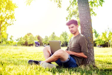çim bir ağacına bakarak veya dizüstü bilgisayar, dışarıda park bir şehir merkezinde çalışan yalan yakışıklı genç adam, öğrenci,