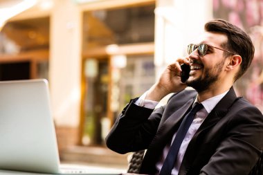 İş adamı güzel üstünde hareket eden telefon, şık takım elbise ve glasesses, dışarıda terasta muhabbet kahve dükkanında oturmuş laptop perde isteyen yakışıklı başarılı adam portresi