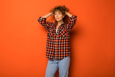 kafasına eller kareli gömlek ve onun holding yanındaki metni için boş alan ile turuncu bir arka plan üzerinde ayakta kot çok kıvırcık kadın