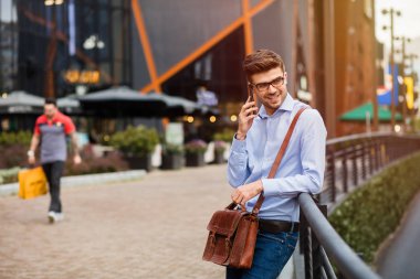 kahve toplantı bekliyor. yakışıklı zeki casual adam gözlük ve deri çanta cep telefonu konuşurken comercial merkezi önünde bir metal çit karşı ayakta dışında