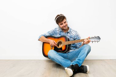 çocuklar için hobi. adam kafasına kulaklık ile gitar çalmak, yere oturmak, onun boş zamanlarında kendi evinde zevk, neden hoşlandığını, beyaz arka plan üzerinde yapıyor