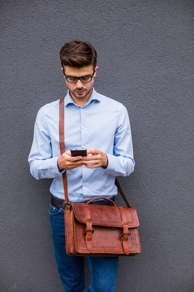 mobile conversation. handsome smart casual man with eyeglasses and leather bag having a conversation on mobile phone on gray abstract wall
