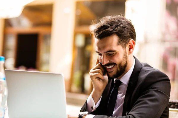 Portrait of handsome successful man with laptop in front, business man having a good conversation at mobile phone and looking to side at terrace in some old town