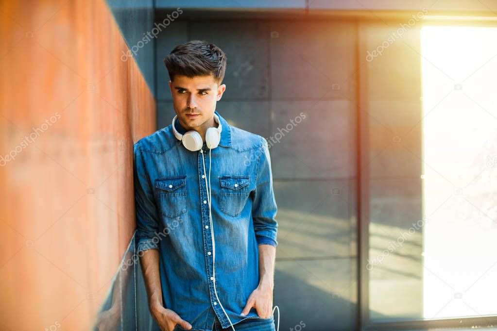 music for youngsters. concept with young guy all in jeans outfit, with headsets around his neck, leaning against a wall