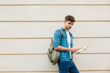 yakışıklı genç adam, bir kitap ve kameraya bakarak bir duvara ayakta dışında sırt çantası ile öğrenci