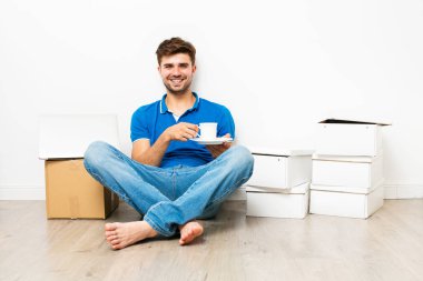 Genç adam bir kahve ya da çay sahip kat onun cardbox yanında, yeni ev için mooving üzerinde mavi gömlekli. Beyaz arka plan üzerinde