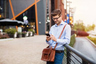 kahve toplantı bekliyor. yakışıklı zeki casual adam gözlük ve comercial merkezi önünde bir metal çit karşı ayakta dışında deri çanta