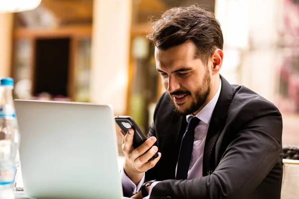 Portrait of handsome successful man outside with laptop in front and looking at his mobile phone in coffee shop, business man having breakfast sitting on beautiful terrace in some old town