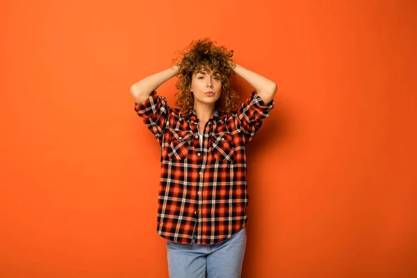 pretty curly woman in a checkered shirt and jeans standing over an orange background with empty space for text next to her holding hands on the head