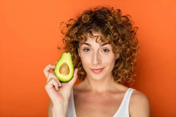young curly woman holding a avocado in hands on orange background