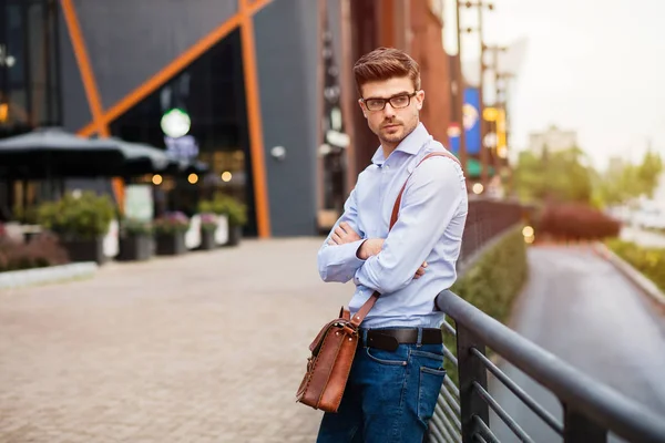 waiting for the coffee meeting.serious handsome smart casual man with eyeglasses and leather bag standing outside against a metal fence in front of comercial center with folded arms