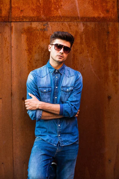 confident guy, cool man in jeans outfit with sunglasses standing against a rusty wall