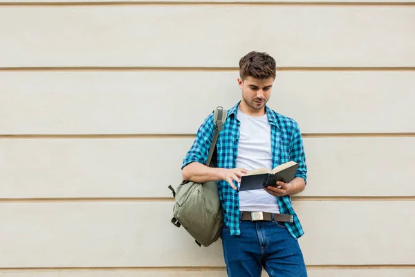 handsome young man ,student with backpack standing outside on against a wall reading a book