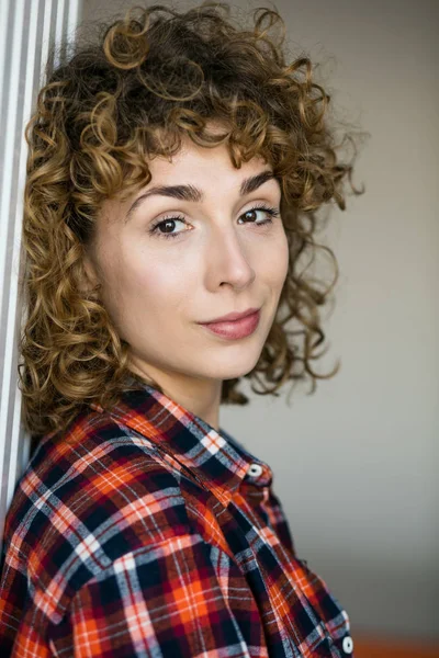 young natural curly woman smiling to camera casual dressed in a checkered shirt inside a house