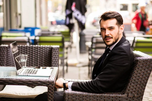 Portrait of handsome successful man with laptop in front looking with confidence at camera, business man having breakfast sitting on beautiful terrace in some old town