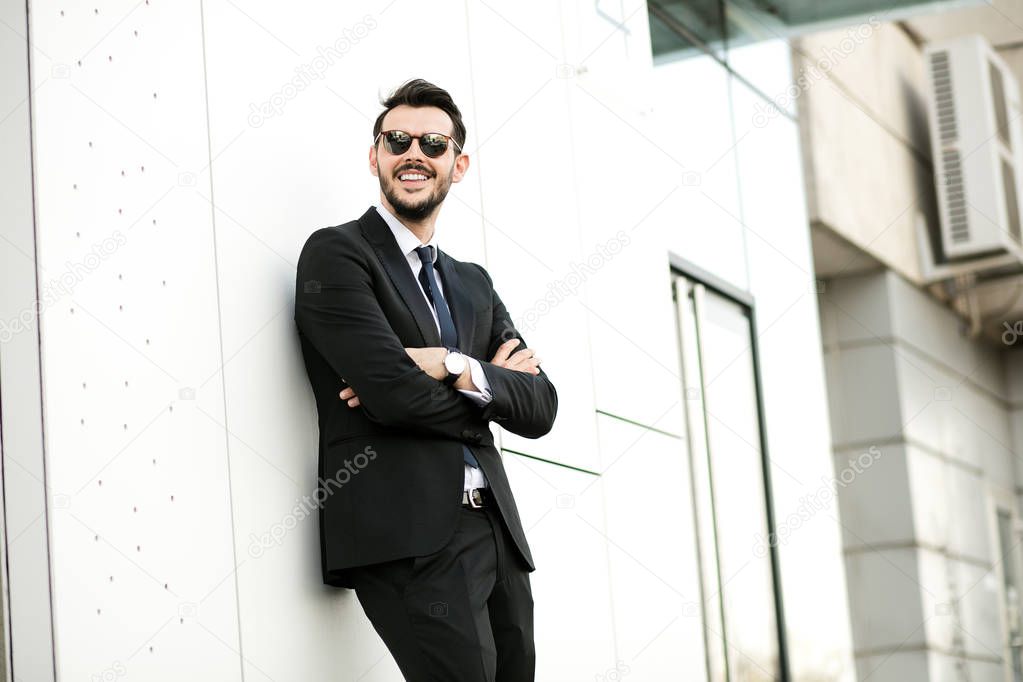 handsome elegant man in suit, businessman, standing outside, waiting for someone, against a white wall with sunglasess