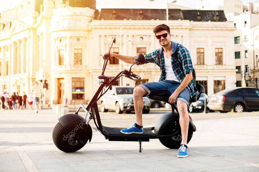 Electric urban transportation. cheerful man sitting with his packpack on electric scooter and smiling to camera in the center of a city