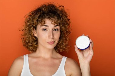 cilt kremi turuncu zemin üzerine küçük bir kutu tutan genç kıvırcık kadın