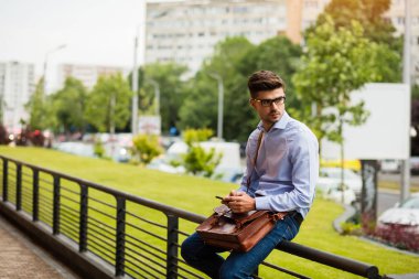 Hala bekliyorum. yakışıklı zeki casual adam gözlük ve metal çit üzerinde duran ve cep telefonunu kullanarak deri çanta