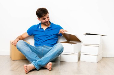 Genç adam oturan kat onun cardbox yanında, yeni ev için mooving üzerinde mavi gömlekli. Beyaz arka plan üzerinde