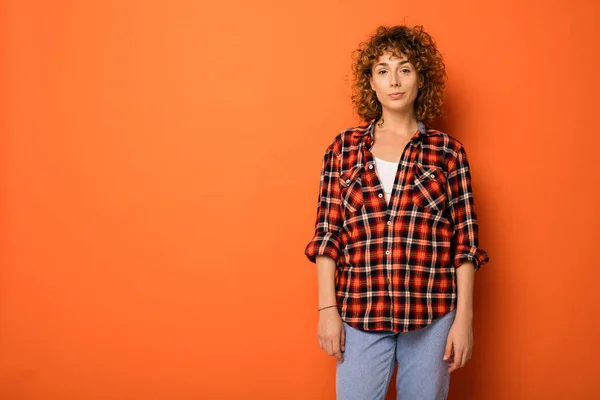 pretty curly woman in a checkered shirt and jeans standing over an orange background with empty space for text next to her