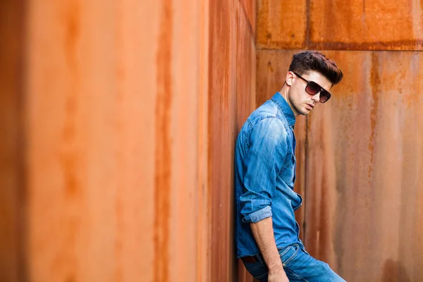 young sexy man in jeans outfit and sunglasses standing against a rusty wall
