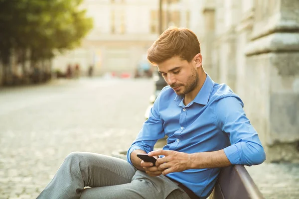 穿着蓝色衬衣的优雅男人 用他的手机在长凳外 — 图库照片