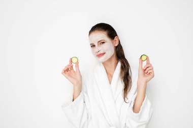 Beyaz arka plan üzerinde yüz maskesi nemlendirici ile güzel genç kız. Bornoz tedaviyi yapıyor ve salatalık dilimleri ile oynarken duş sonra kız fotoğrafı. Cilt bakım kavramı