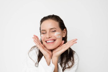 Genç kadın portresi krem cilt tedavisi için fotoğraf makinesi ile sevinç ve zevk gülümseyen yüzünde iki satır ile. sonra onu bornoz duşta Islak saçla