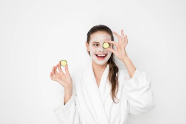 Beyaz arka plan üzerinde yüz maskesi nemlendirici ile güzel genç kız. Bornoz tedaviyi yapıyor ve salatalık dilimleri ile oynarken duş sonra kız fotoğrafı. Cilt bakım kavramı