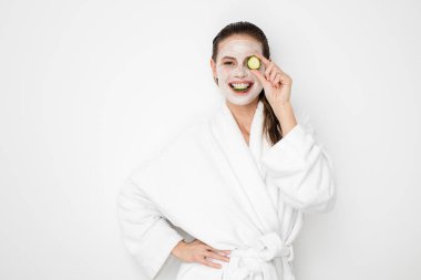 Beyaz arka plan üzerinde yüz maskesi nemlendirici ile güzel genç kız. Bornoz tedaviyi yapıyor ve salatalık dilimleri ile oynarken duş sonra kız fotoğrafı. Cilt bakım kavramı