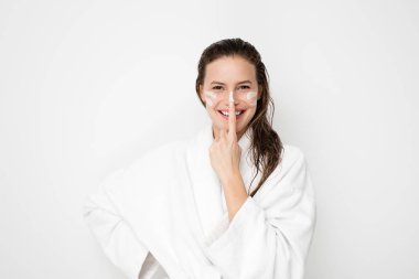 Genç kadın kusursuz bir cilt, yüzünde nemlendirici krem uygulamak. Krema ile oynama ve yüzündeki çizgiler yapmak. Sonra banyo beyaz bornoz ve havlu beyaz arka plan üzerinde kadın fotoğrafı. Cilt bakım kavramı