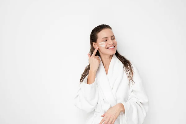 Young woman with flawless skin, applying moisturizing cream on her face. Photo of woman after bath in white bathrobe and towel on white background. Skin care concept