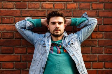 boyun arkasında ellerini tutarak ve kamera, bir tuğla tüm ayakta isteyen genç rahat adam kot ceketli
