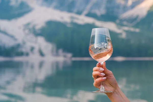 Braies Gölü kıyısında bir bardak şarap çeviren genç bir bayanın eli. Arka planda yeşil bir orman var.