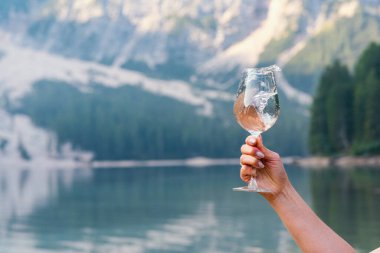 Braies Gölü kıyısında bir bardak şarap çeviren genç bir bayanın eli. Arka planda yeşil bir orman var.